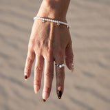 A delicate pearl beaded bracelet with small silver charms, on a model's wrist with sand background.