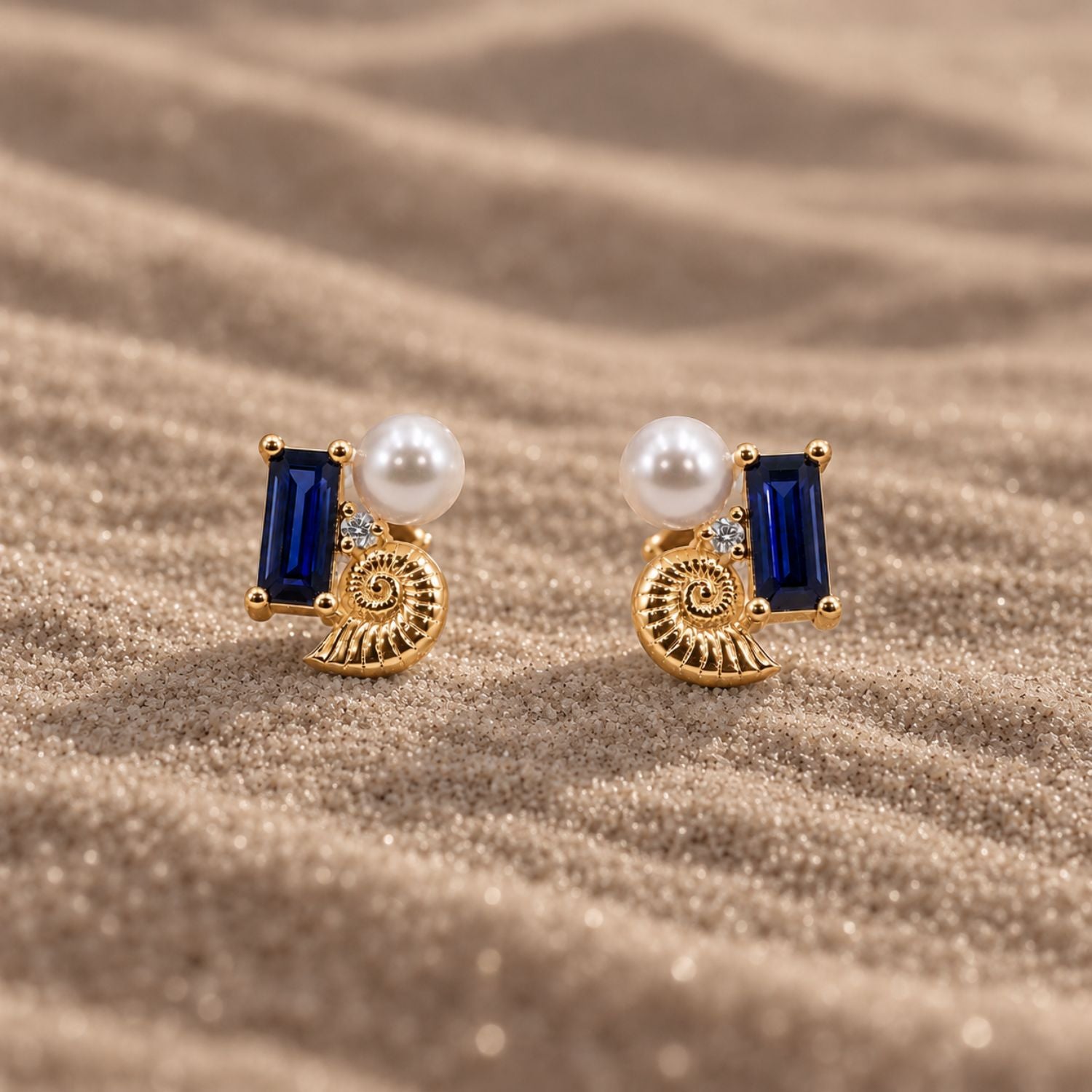 14K gold vermeil stud earrings with a blue sapphire, shell, lab-grown diamond and pearl on sand.