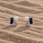 Sterling silver stud earrings with a blue sapphire, shell, lab-grown diamond and pearl on sand.