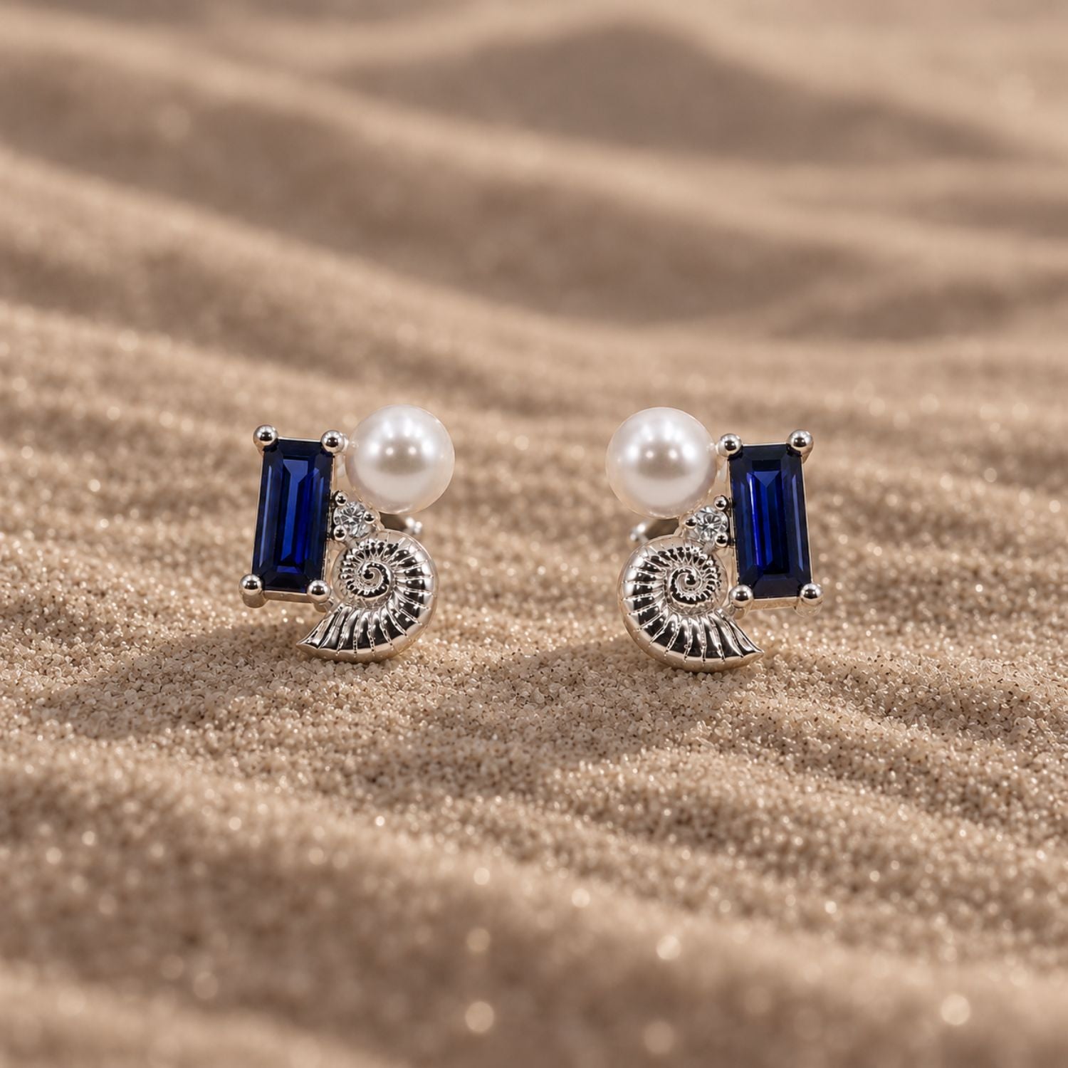 Sterling silver stud earrings with a blue sapphire, shell, lab-grown diamond and pearl on sand.