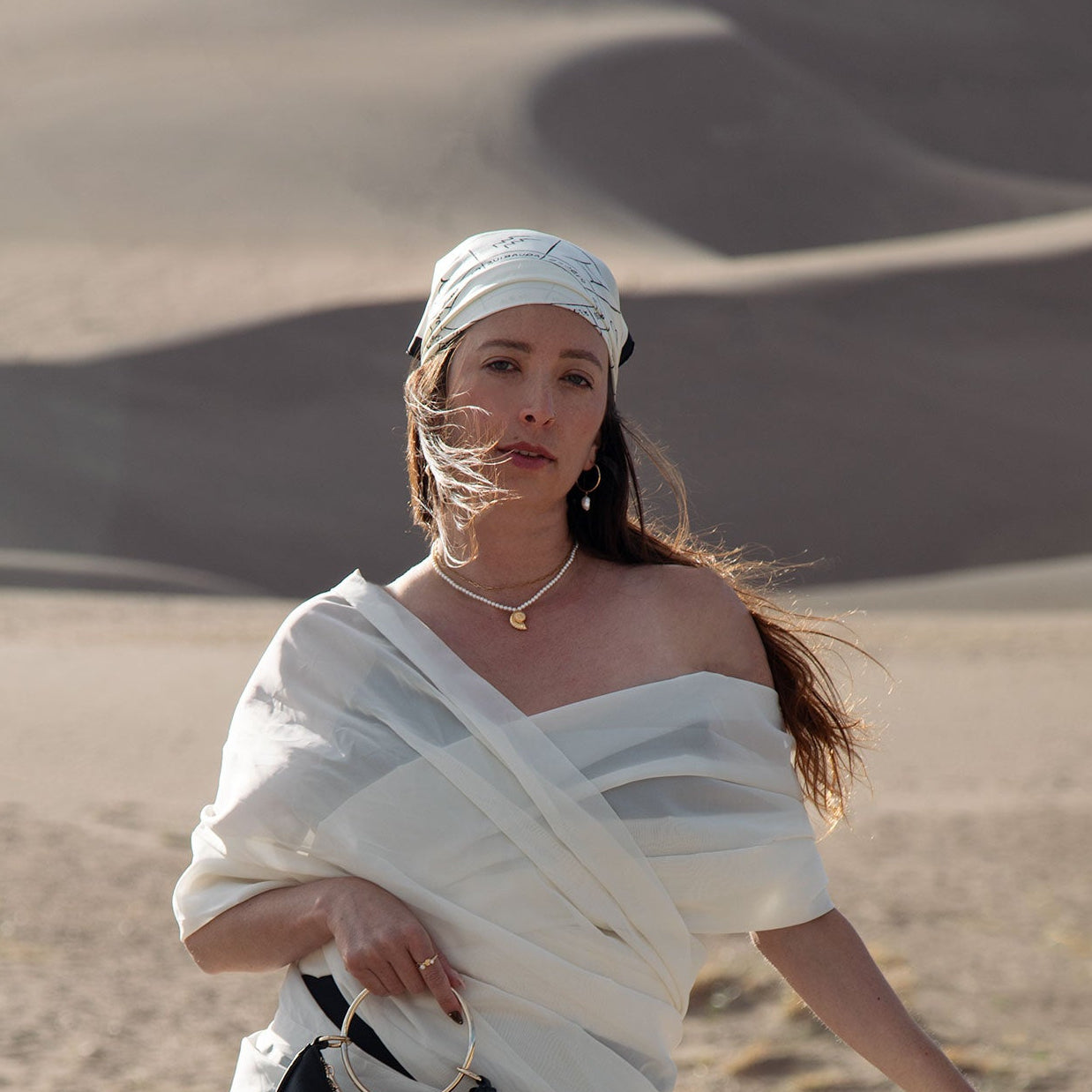 A girl in a desert wearing a pearl shell necklace, scarf around her head and white fabric draped around her body with her hair blowing in the wind. 