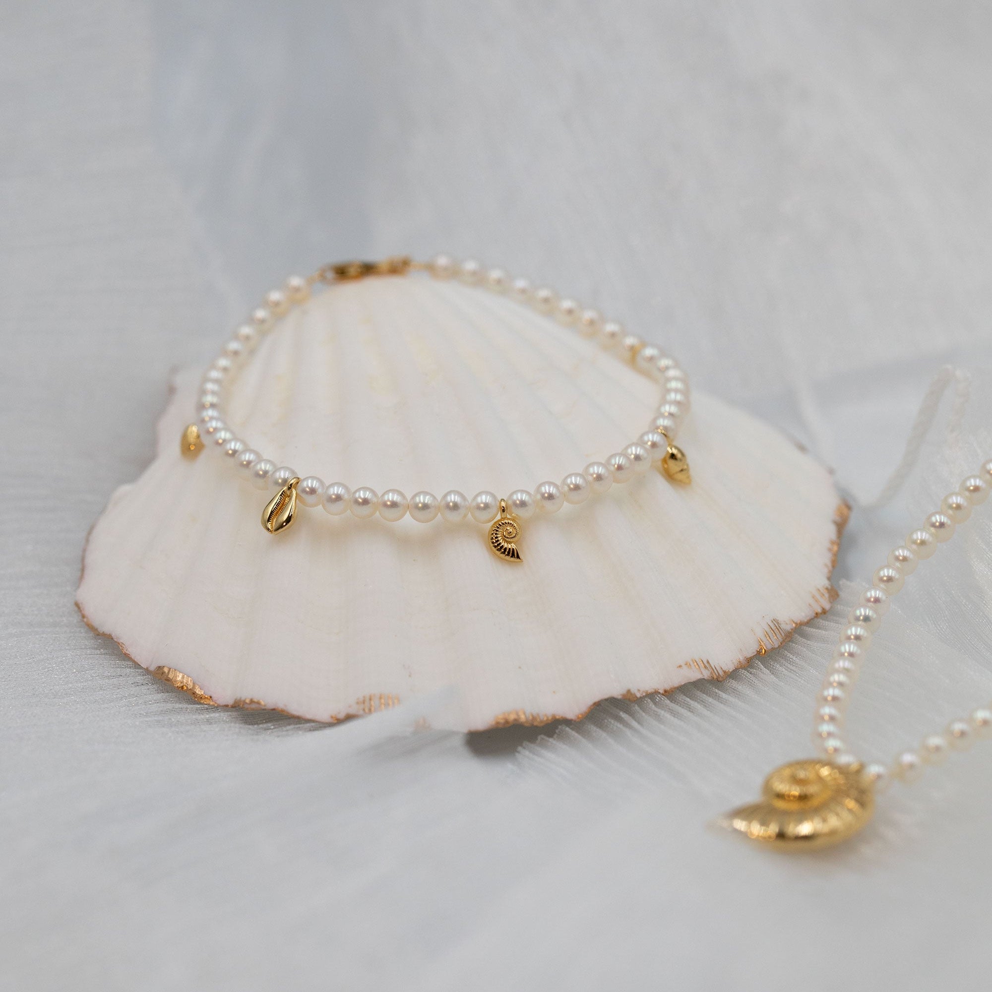 A delicate pearl bracelet with small gold shell charms is displayed on a white seashell, with a matching gold necklace partially visible on the right, set against a soft white fabric background.
