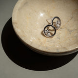 A pair of silver-toned hoop earrings with rectangular and pear lab-grown diamonds, resting inside a shallow beige stone bowl on a neutral surface.