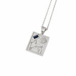 A silver-toned rectangular pendant necklace featuring an engraved constellation design, a small textured circular detail, and a dark blue rectangular inset near the top left, suspended from a matching chain on a white background.