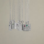 Eight silver pendant necklaces with detailed zodiac engravings and small colored gemstone accents, hanging against a solid beige background.