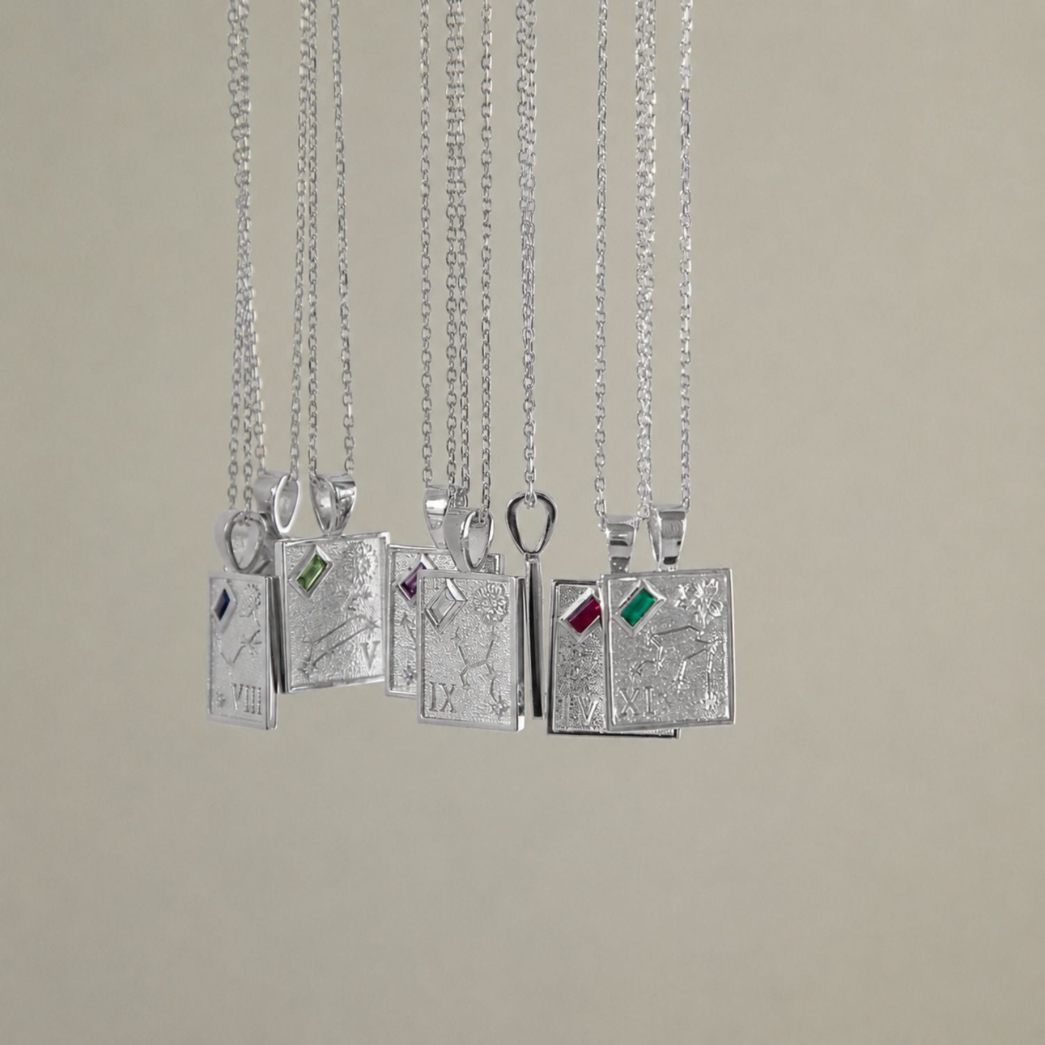 Eight silver pendant necklaces with detailed zodiac engravings and small colored gemstone accents, hanging against a solid beige background.