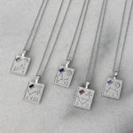 Five silver pendant necklaces with engraved zodiac constellations and small colored gemstone accents, arranged diagonally on a light gray marble surface.