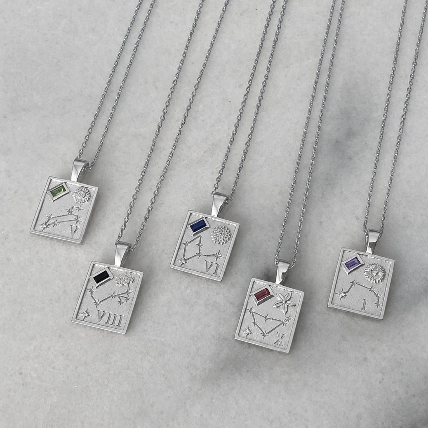 Five silver pendant necklaces with engraved zodiac constellations and small colored gemstone accents, arranged diagonally on a light gray marble surface.