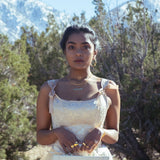 A model wearing gold jewelry and holding a small bag. She is wearing a satin pink dress and there are mountains and greenery in the background. 