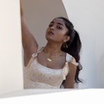 A model wearing gold jewelry and framed by a white wall in front, and a white archway behind her. She is wearing a satin pink dress with bows near the shoulders. 