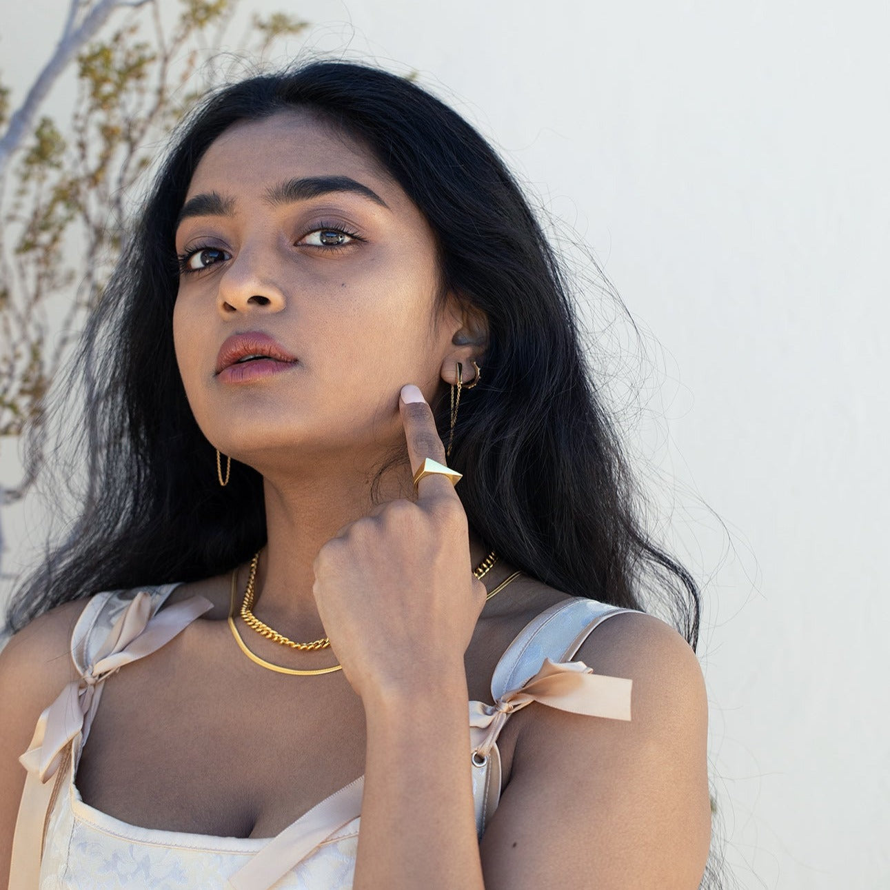 A model posing with gold jewelry. She's wearing a pin satin dress with a small brand behind her and white background.