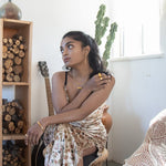 A model in a rustic setting with chopped wood on a shelf , and a guitar behind her. She's wearing a dress and gold vermeil jewelry.