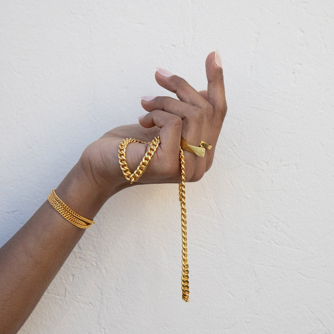 A gold geometric ring with a small square diamond on a model's hand with other gold jewelry, with a white background. 