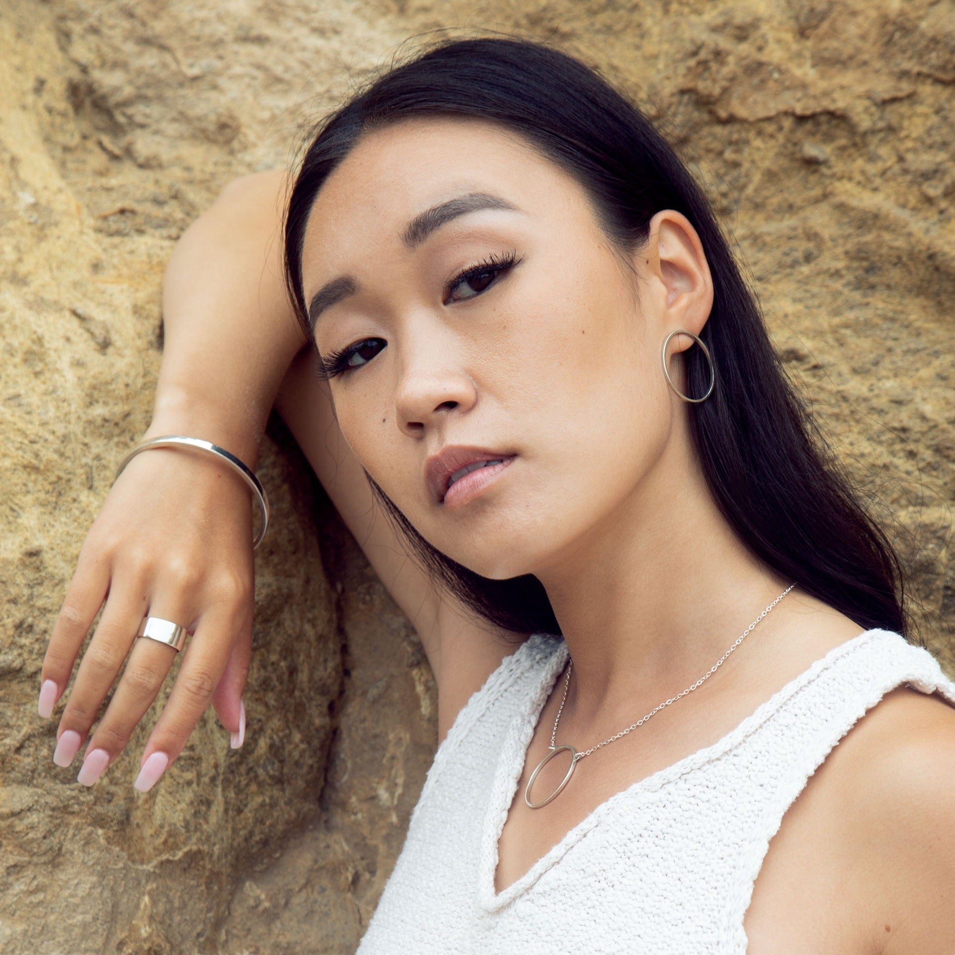 A model posing against a rock wearing a sterling silver necklace, earrings, bracelet, and ring. She is wearing a white knit top. 