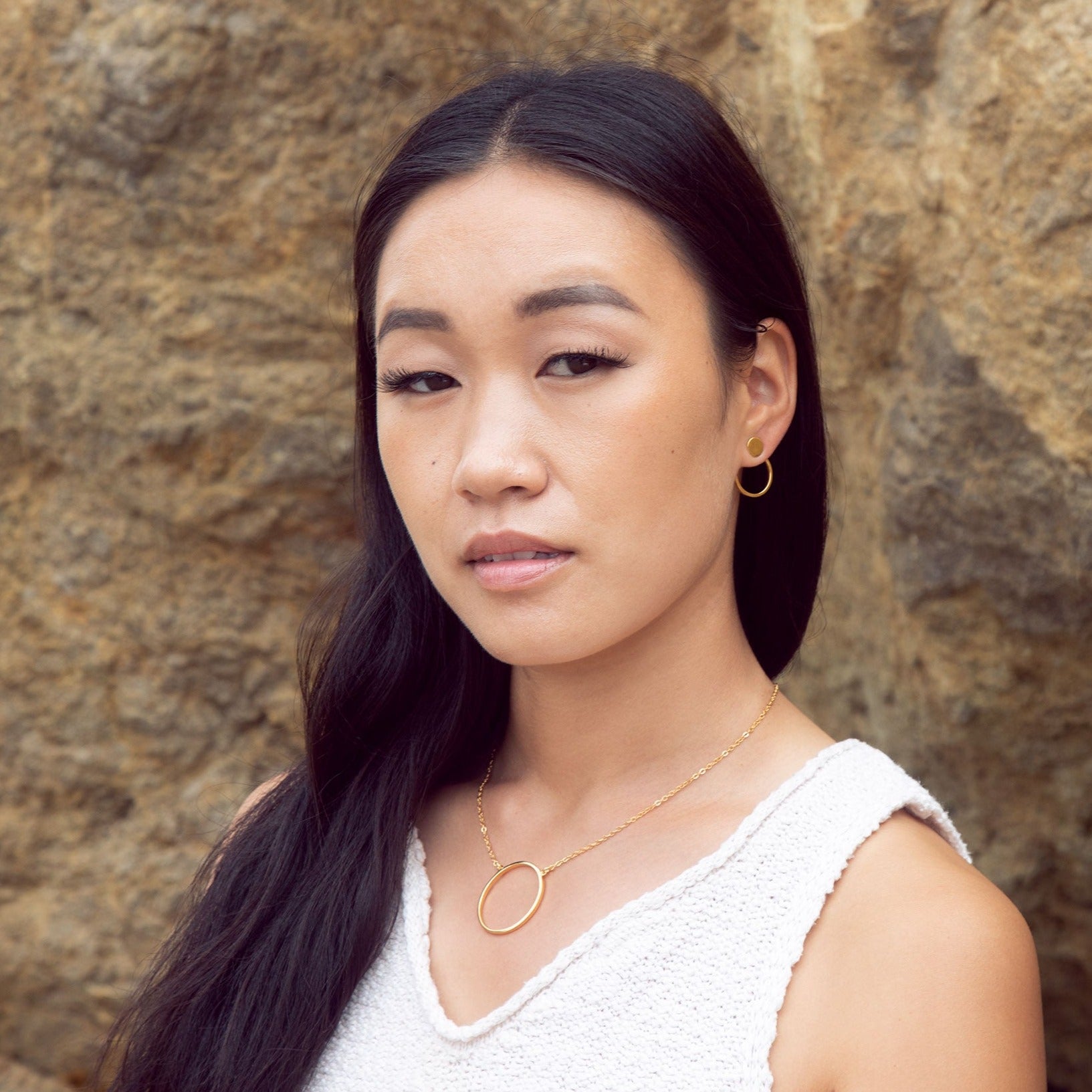 A 14K gold plated circular necklace on a model who is wearing a white shirt and gold earrings posing in front of a rocky background. 