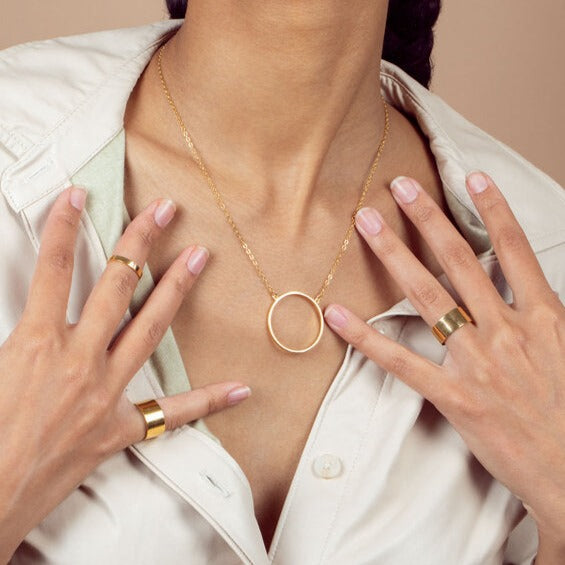 A 14K gold plated circular necklace on around a model's neck with her hands on her chest featuring gold rings, shot in studio with a tan background.