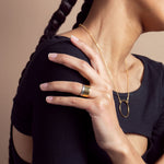 A 14K gold plated circular necklace con a model who is wearing a black shirt and wearing two toned rings on her ring finger. Shot in studio on a tan background.