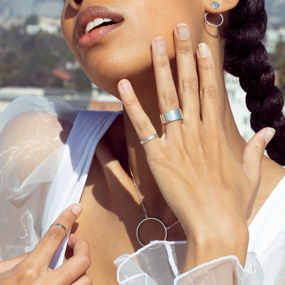 A cyclical thick ring made of sterling silver and a thin ring on a model's hand who is touching her face. She is also wearing a silver necklace, earrings, and white top, posing outside. 