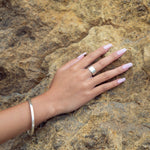 A cyclical thick ring made of sterling silver, displayed on a model's finger who has long pink nails and is wearing a sterling silver bangle, set on a rocky background.