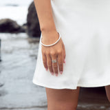 A cyclical thick ring made of sterling silver on a model's hand who also has a sterling silver bangle. She is wearing a white dress and posing at the beach. 