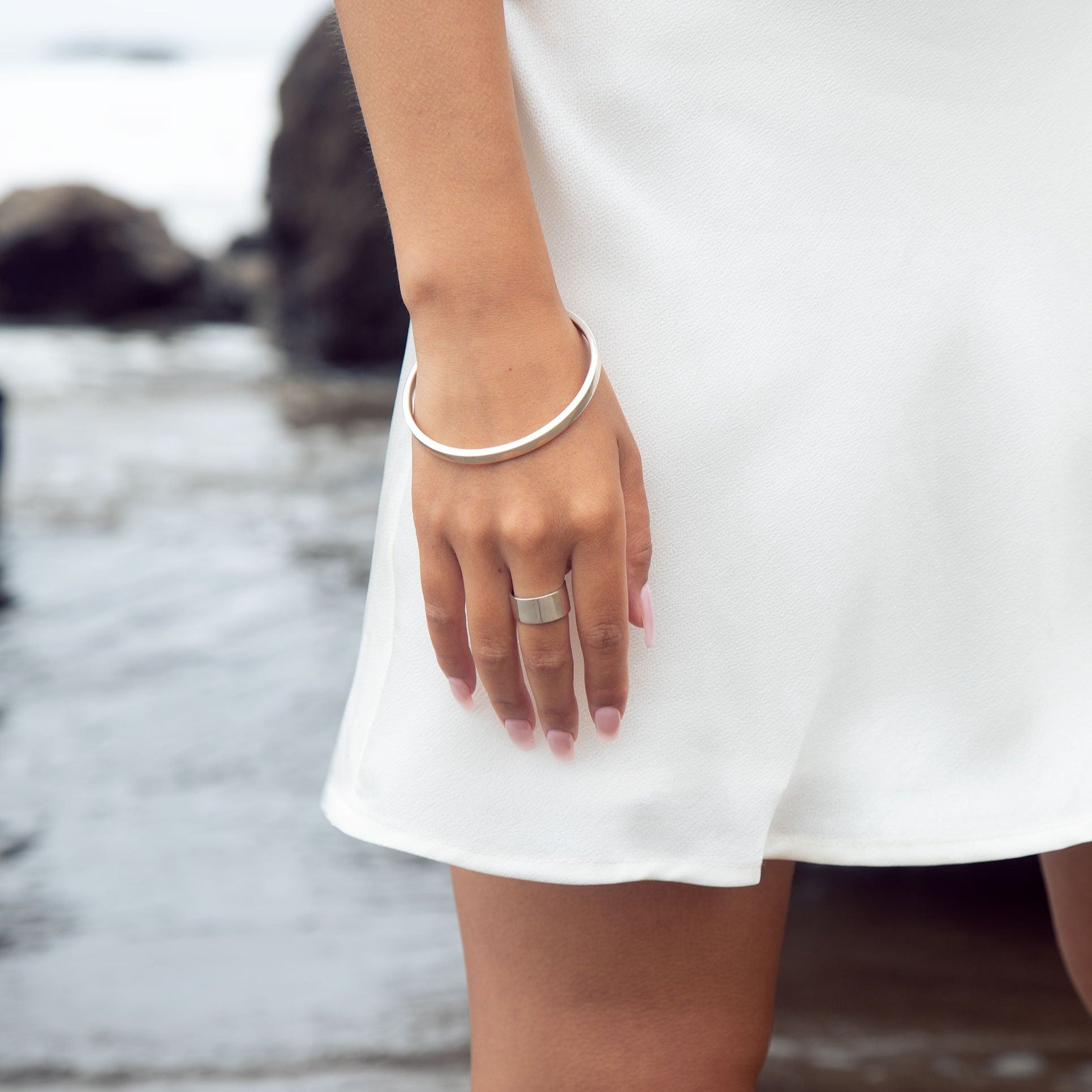 A cyclical thick ring made of sterling silver on a model's hand who also has a sterling silver bangle. She is wearing a white dress and posing at the beach. 