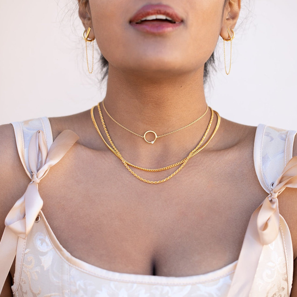 A model wearing three gold necklaces and gold earrings with a white wall in the background. She has on a pink top with bows near the shoulders. 