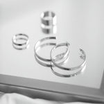 A black and white photo featuring a pair of polished silver-tone open hoop earrings on a plain white background with two rings in the background.