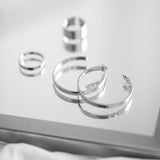 A black and white photo featuring a pair of polished silver-tone open hoop earrings on a plain white background with two rings in the background.