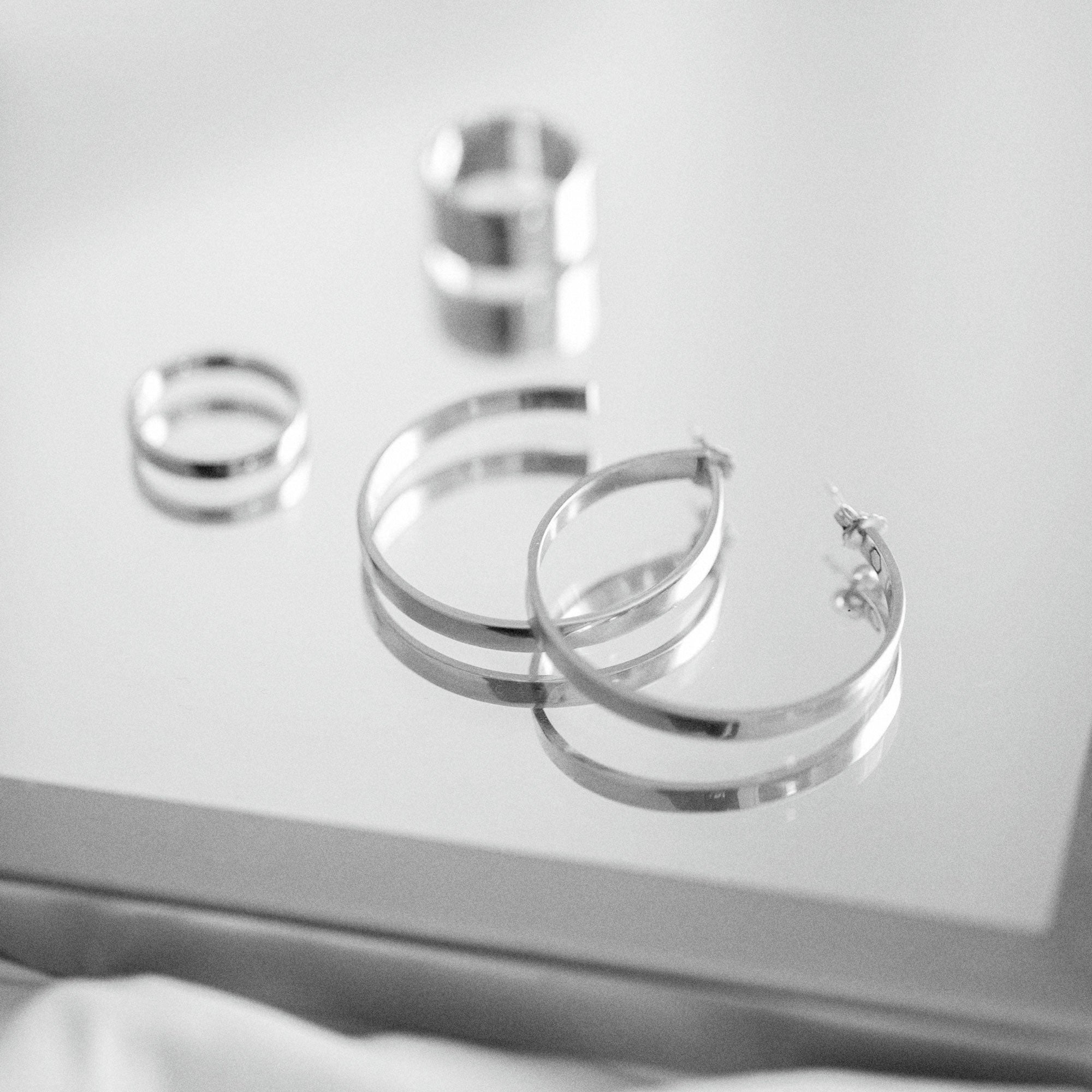 A black and white photo featuring a pair of polished silver-tone open hoop earrings on a plain white background with two rings in the background.