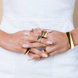 Gold and rose gold toned chunky rings on a model's intertwined fingers. She is wearing a white dress in a studio. 