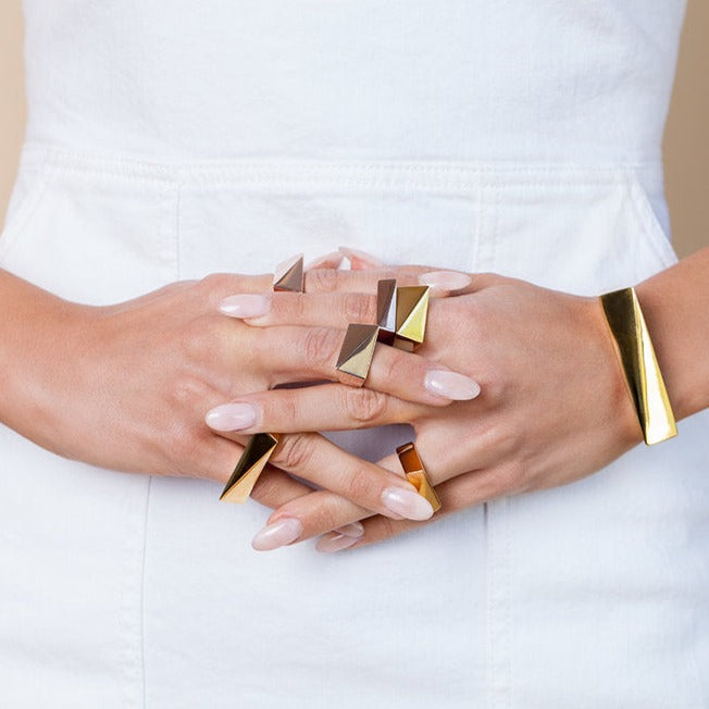 Gold and rose gold toned chunky rings on a model's intertwined fingers. She is wearing a white dress in a studio. 