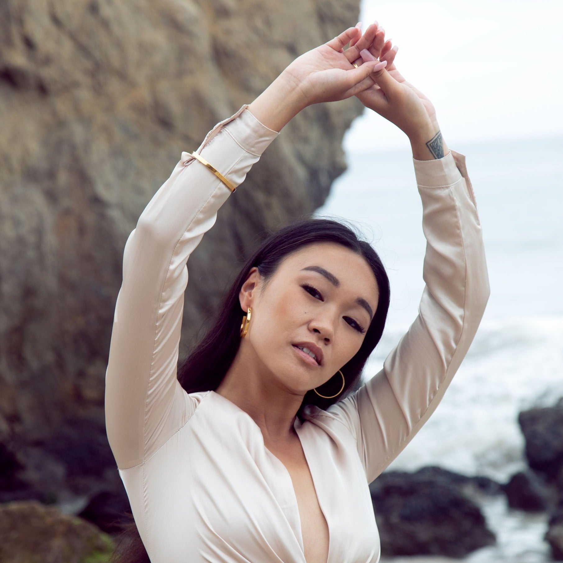A pair of gold vermeil hoop earrings on a model who is wearing a satin pink dress and posing at the beach with rock in the background. 