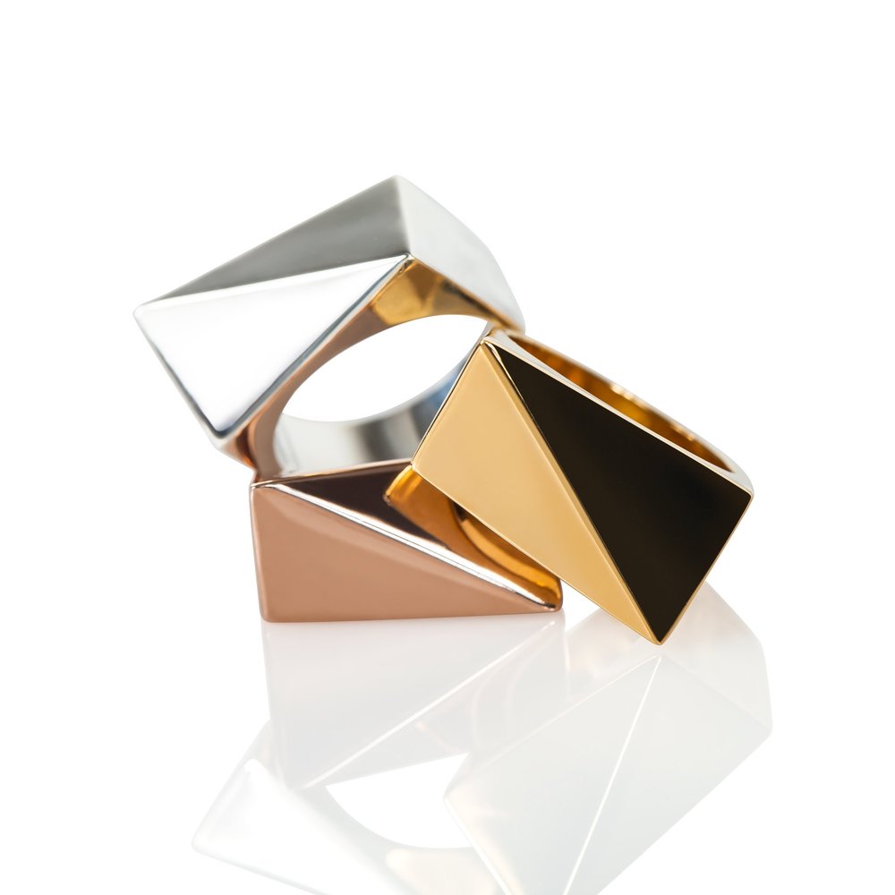 Three chunky gold, rose gold, and sterling silver statement rings stacked on a white background with a reflection below them. 