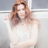 A redheaded model in a silver jumpsuit wearing a 14K gold vermeil necklace with a geometric slanted rectangular pendant standing in front of a silver air stream. 