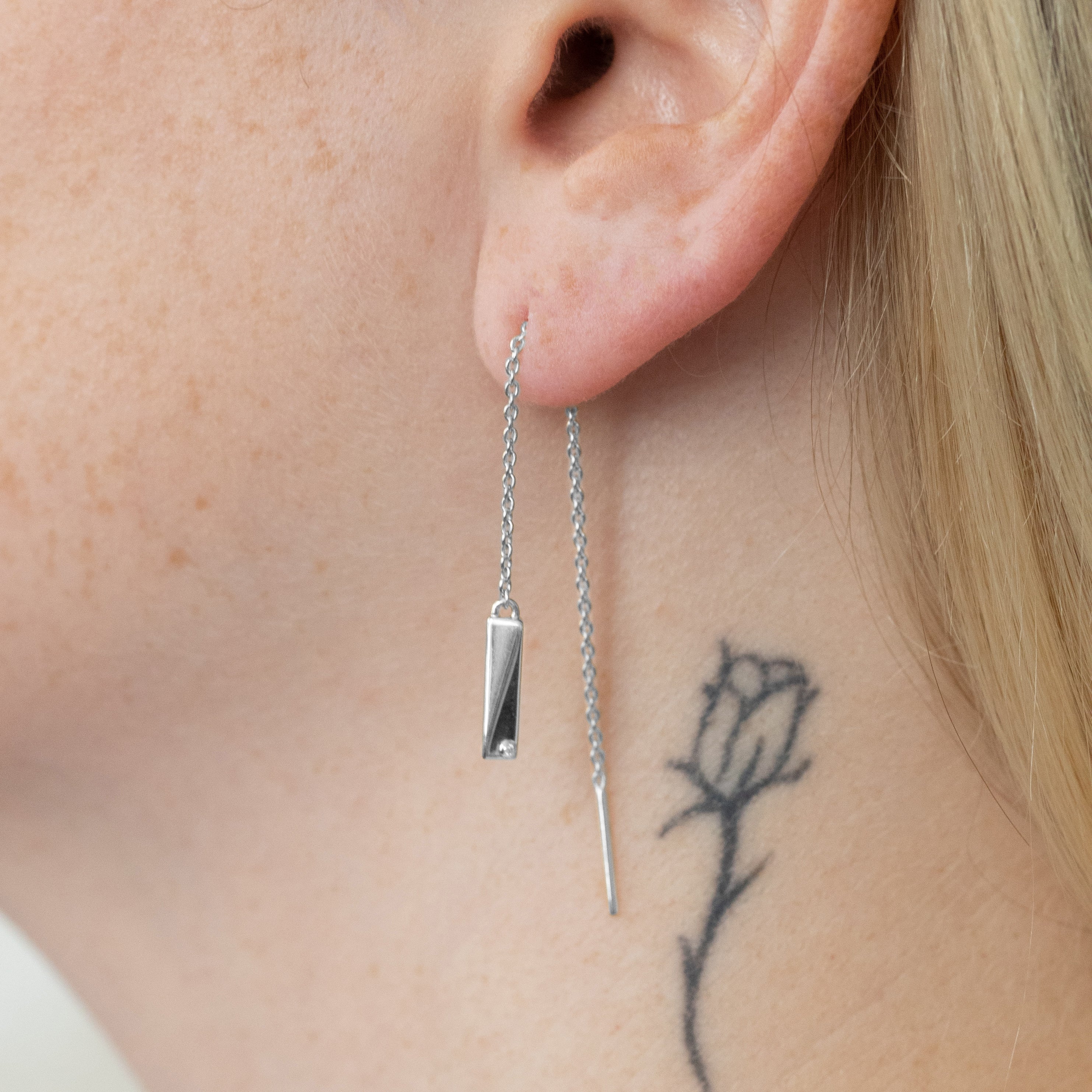 A pair of silver drop earrings with long chain, rectangular bar pendants, and slim dangling pendants with lab-grown diamonds, close up on a model's ear. 