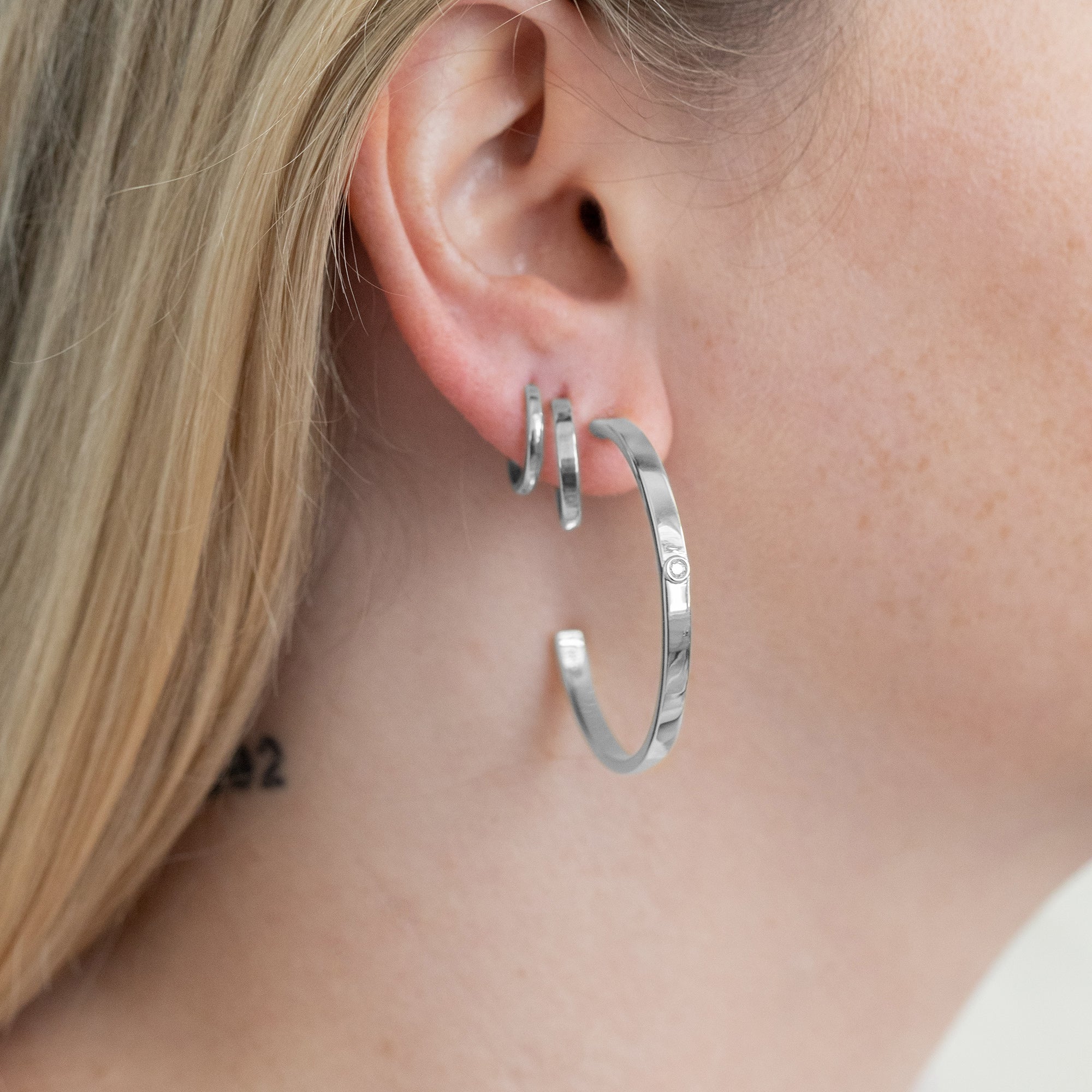 A close up of a model's ear with a large silver hoop with lab-grown diamond on the right side and two smaller hoops in sterling silver in the second and third piercings.